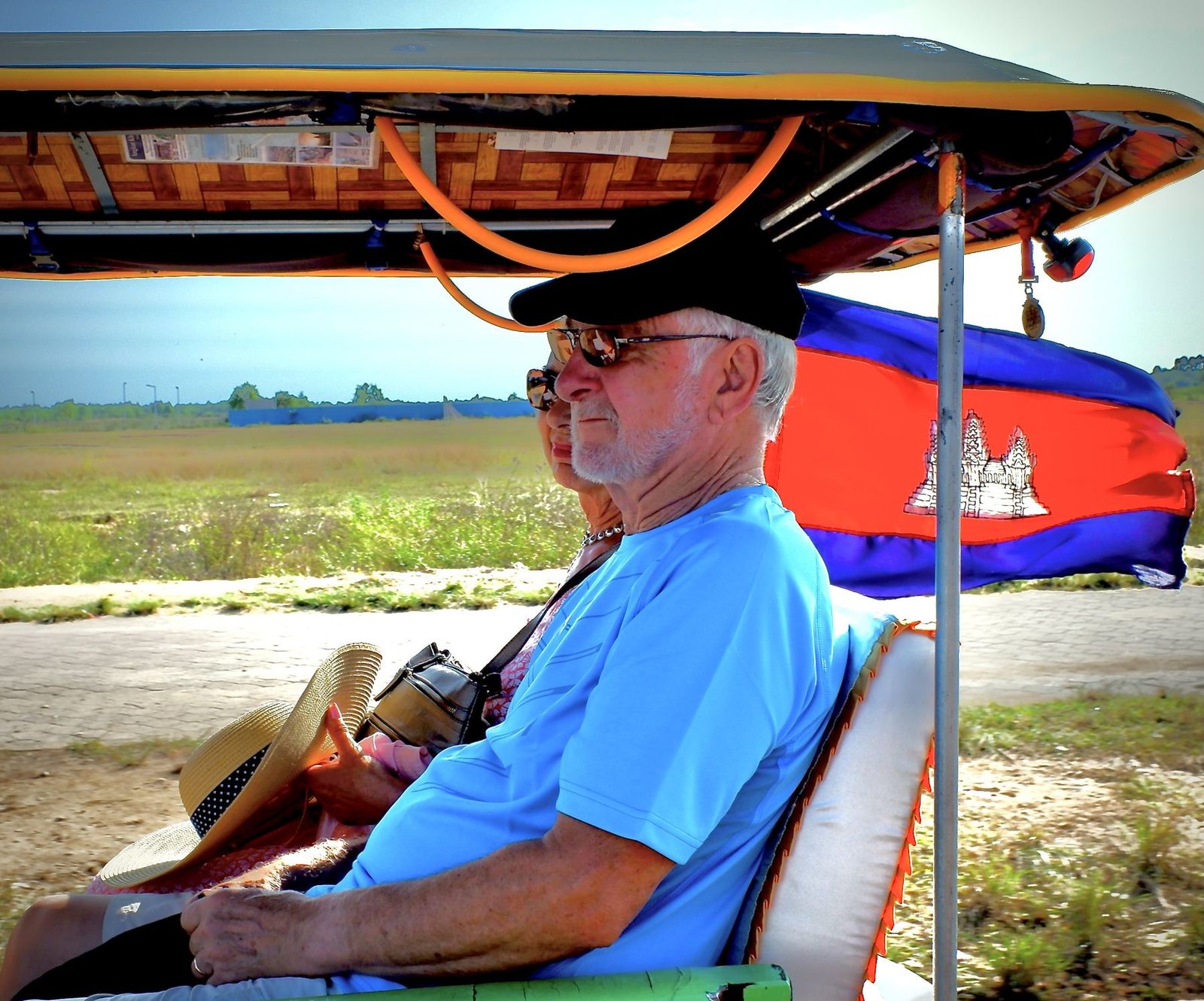A man rides in the back of an open-air tuk tuk, Cambodian flag flying beside him