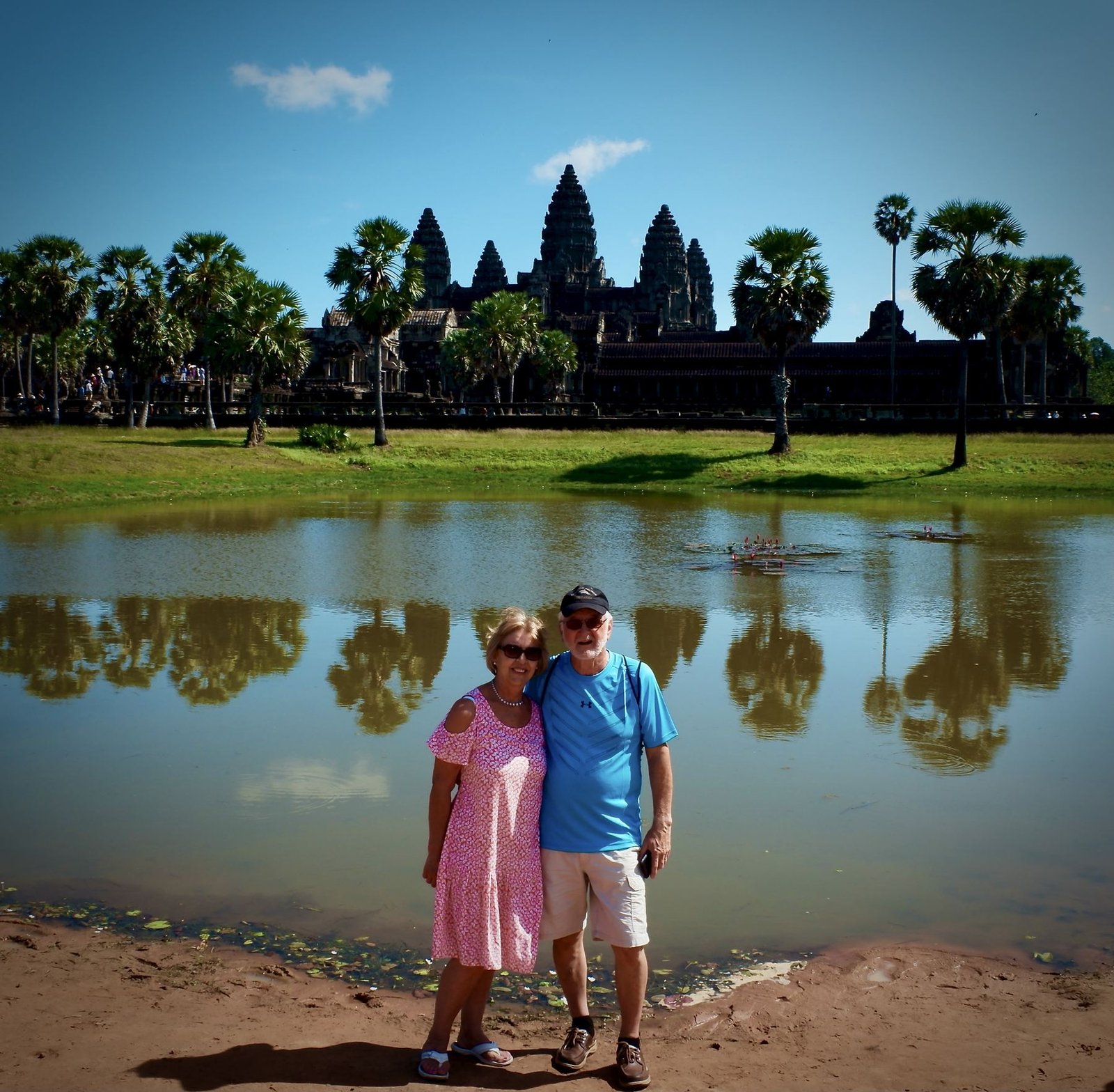 A couple stands in front of Angkor Wat, its towers reflected in the moat behind them