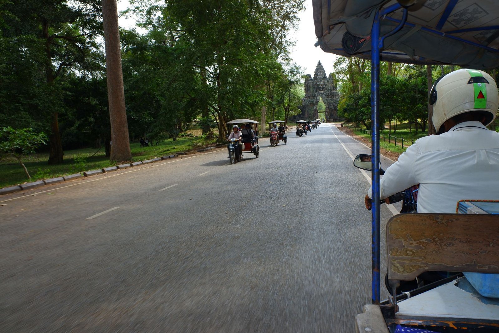 View from a moving tuk tuk down a tree-lined road, an ancient stone gate visible in the distance
